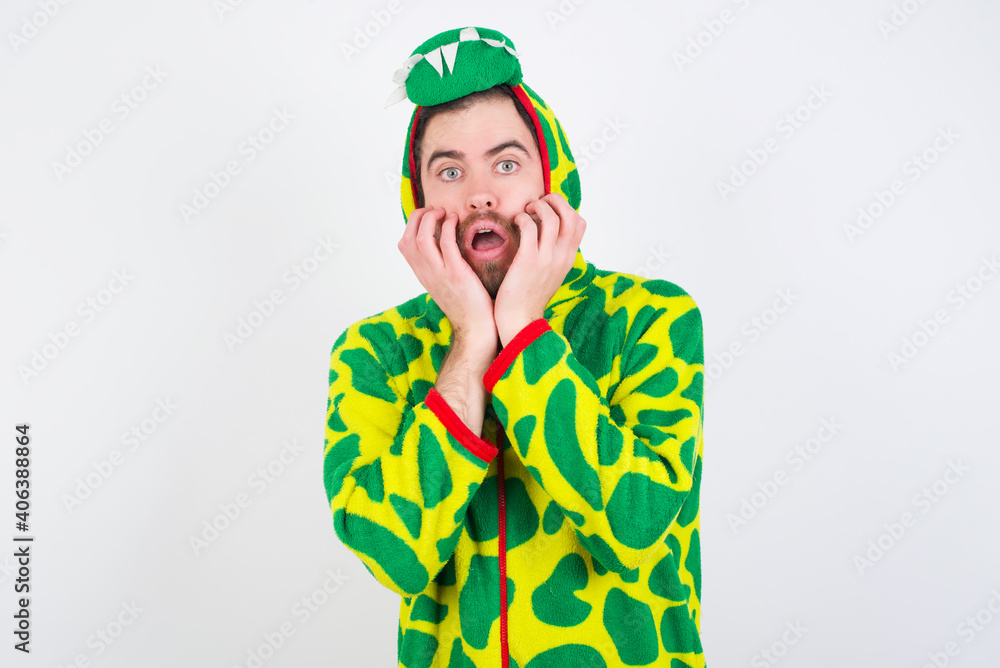Fototapeta premium Stupefied Young caucasian man wearing a pajama standing against white background expresses excitement and thrill, keeps jaw dropped, hands on cheeks, has eyes popped out