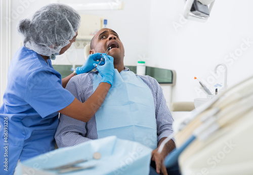 Hispanic man sitting in dental chair  getting anesthetic injection before teeth treatment by professional female dentist