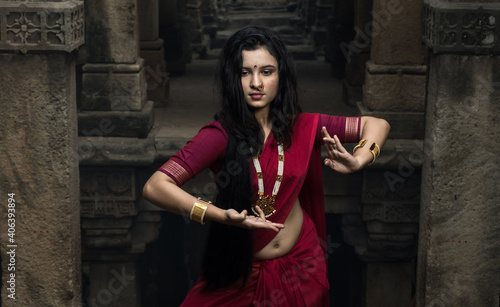 Portrait of beautiful Indian girl in heritage stepwell wearing traditional Indian red saree, gold jewellery and bangles holding religious plate. Maa Durga agomoni shoot concept