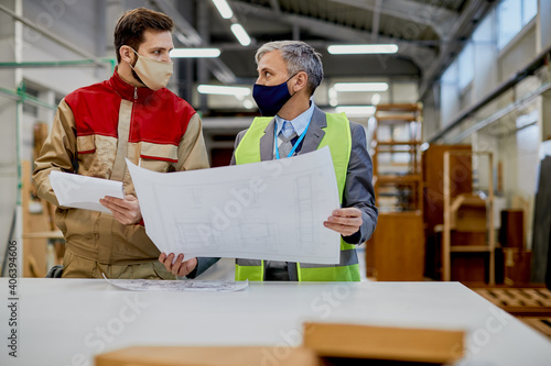 Quality control inspector and woodworking laborer wearing face masks while examining design plans. © Drazen
