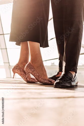 Close up a woman in elegant black dress and rose golden high heels staying together with her man in black pants and shoes. Elegant couple staying in front of sunlight on wooden floor near railings