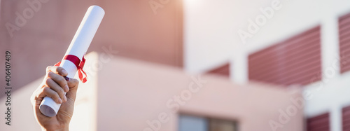Close-up shot of a university graduate holding degree certification to shows and celebrate his success in the college commencement day with sunlight in the background.