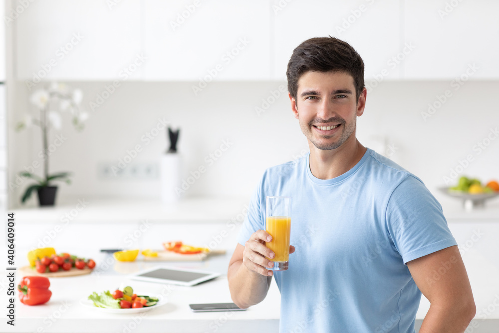 Handsome man in a blue T-shirt with a glass of orange juice in his hands in the kitchen at home Handsome man in a blue T-shirt with a glass of orange juice in his hands in the kitchen at home