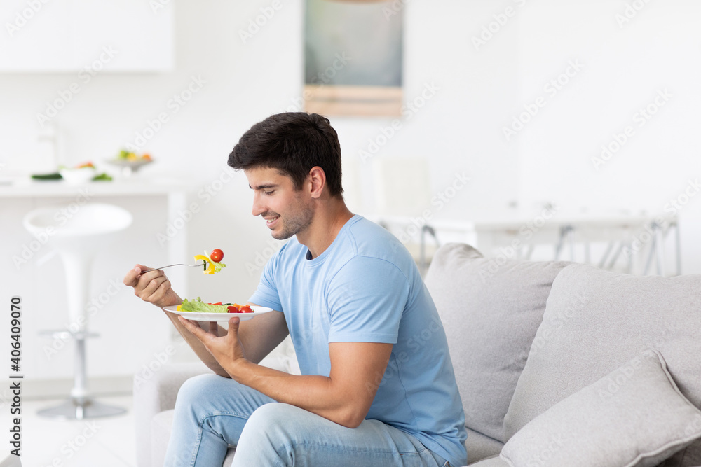 Handsome man eating fresh vegetable salad while sitting on the couch at home, healthy and vegan food