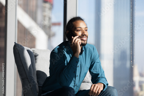 Smiling young African American man sit on chair in office or home talk on smartphone with colleague or client. Happy ethnic male relax speak have cellphone call using good mobile provider connection.