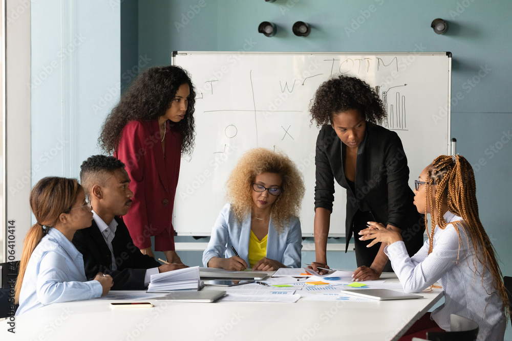 Obraz premium Focused multiethnic diverse colleagues coworkers gather at desk in office boardroom discuss company paperwork together. Multiracial businesspeople talk brainstorm at team meeting at workplace.