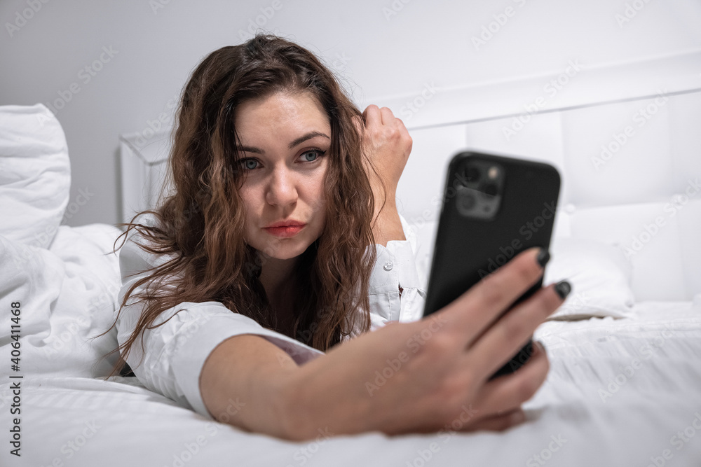 Attractive young woman with long wet hair making selfie with contemporary smartphone lying on comfortable large bed at home Attractive young woman with long wet hair making selfie with contemporary smartphone lying on comfortable large bed at home
