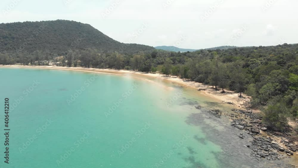 Rocky golden sandy shoreline with lush green hills encircling the shallow turquoise waters of M'pai Bay, Koh Rong Sanloem, Cambodia - Aerial low angle fly-over shot