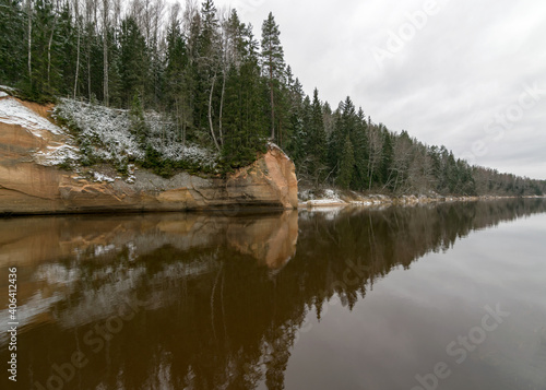 Sandstone rock wall by the river with forest, rock reflection in the river water, View of the Gauja river in December with light snow and cloudy sky, Gauja National Park photo