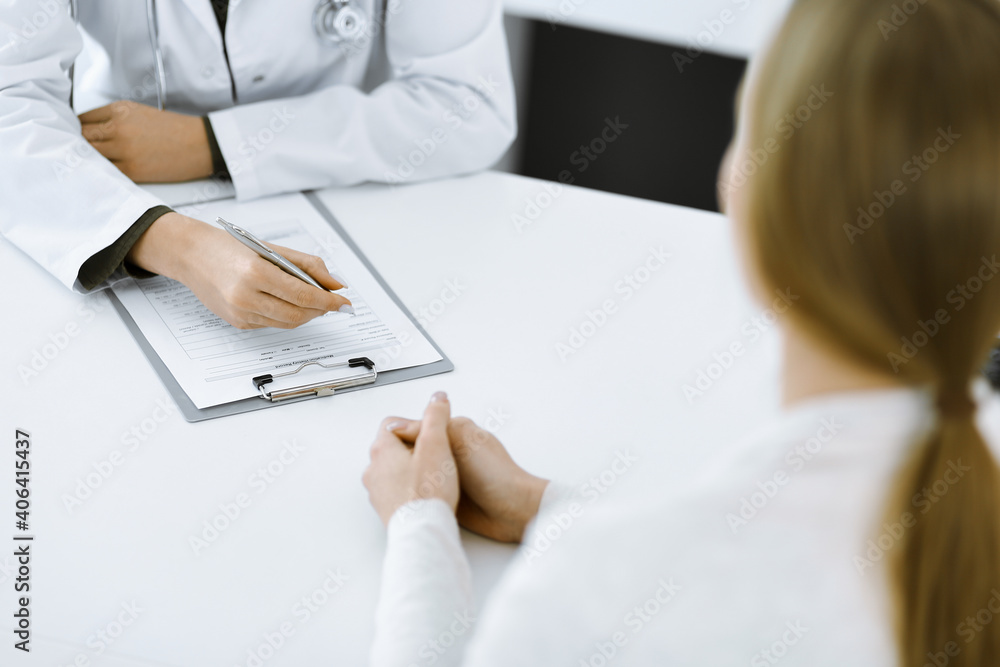 Unknown woman-doctor and female patient sitting and talking at medical examination in clinic, close-up. Therapist wearing green blouse is filling up medication history record. Medicine concept Unknown woman-doctor and female patient sitting and talking at medical examination in clinic, close-up. Therapist wearing green blouse is filling up medication history record. Medicine concept