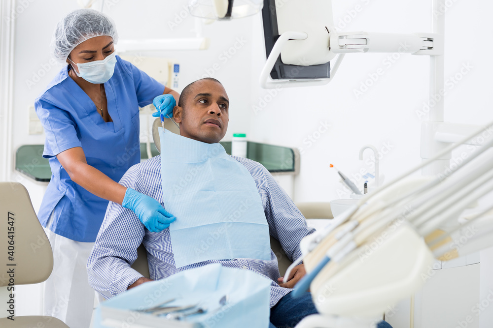 Obraz premium Portrait of adult hispanic man sitting on dental chair while woman dentist preparing him for medical examination in clinic