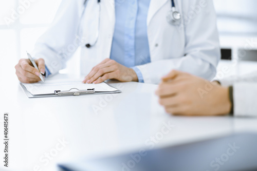 Unknown woman-doctor and female patient sitting and talking at medical examination in clinic  close-up. Therapist wearing blue blouse is filling up medication history record. Medicine concept