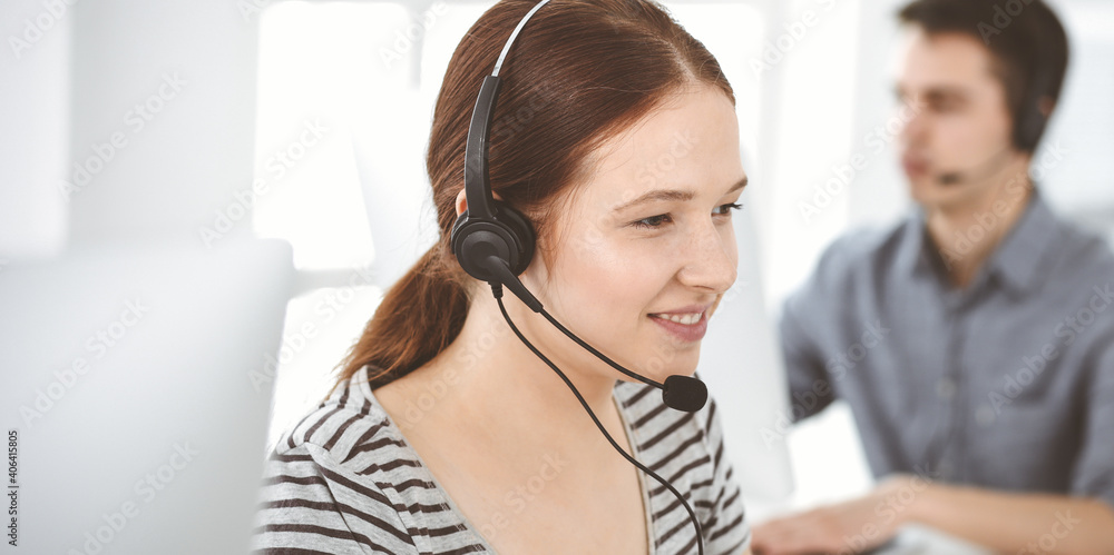 Naklejka premium Casual dressed young woman using headset and computer while talking with customers online. Group of operators at work. Call center, business concept
