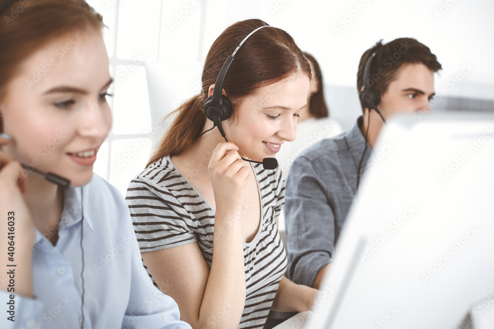 Casual dressed young woman using headset and computer while talking with customers online. Group of operators at work. Call center, business concept Casual dressed young woman using headset and computer while talking with customers online. Group of operators at work. Call center, business concept