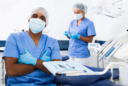 Portrait of qualified latin american dentist standing in blue uniform in modern medical dental office