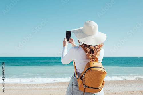 Woman using her smartphone wearing a hat and a yellow backpack bag at the beach