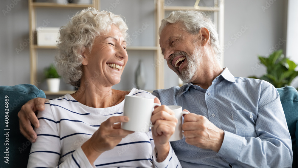Overjoyed middle aged retired married couple relaxing on comfortable sofa, enjoying communicating while drinking hot morning coffee together at home, laughing at funny joke, family relations concept. - obrazy, fototapety, plakaty 