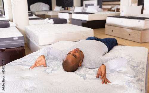 Smiling adult hispanic man lying on bed in furniture store, enjoying convenience of modern orthopedic mattress .