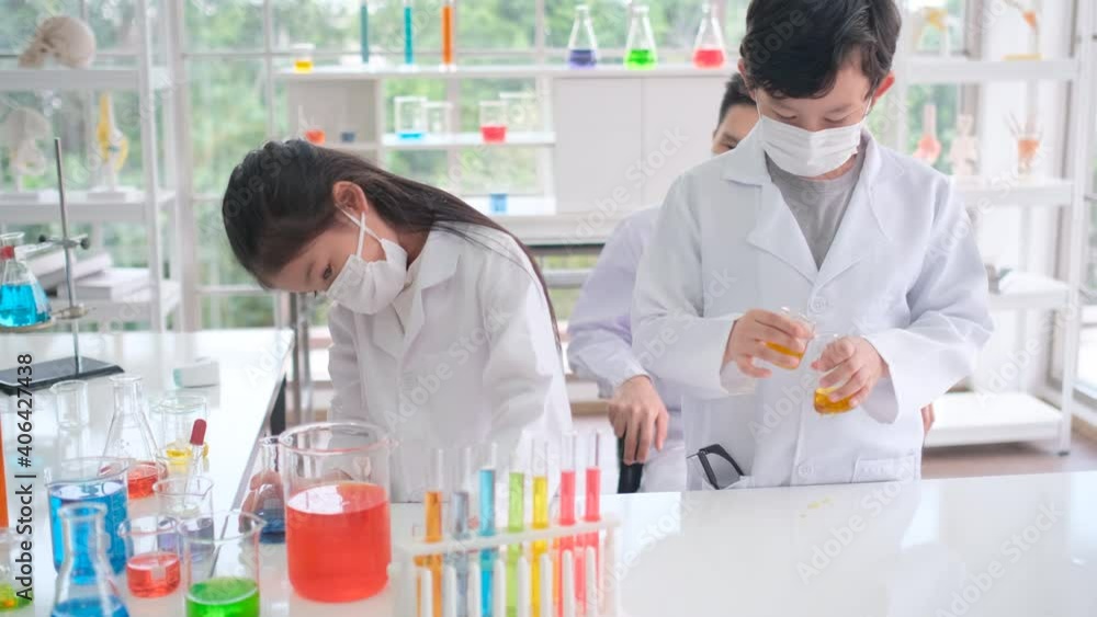 Young scientist girl and boy with hygiene mask enjoy with testing of colorful chemical experiment in laboratory or classroom and teacher look after in the back of room. Young scientist girl and boy with hygiene mask enjoy with testing of colorful chemical experiment in laboratory or classroom and teacher look after in the back of room.