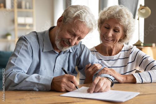 Happy old mature retired family couple putting signature on paper document, feeling satisfied with purchasing own apartment house or signing medical insurance agreement together at office meeting.
