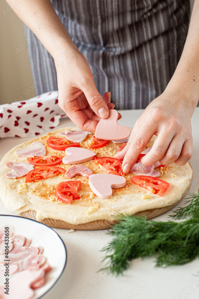 The process of making homemade pizza for Valentine's Day. Women's hands lay the ingredients cut in the form of hearts on the base of the dough.