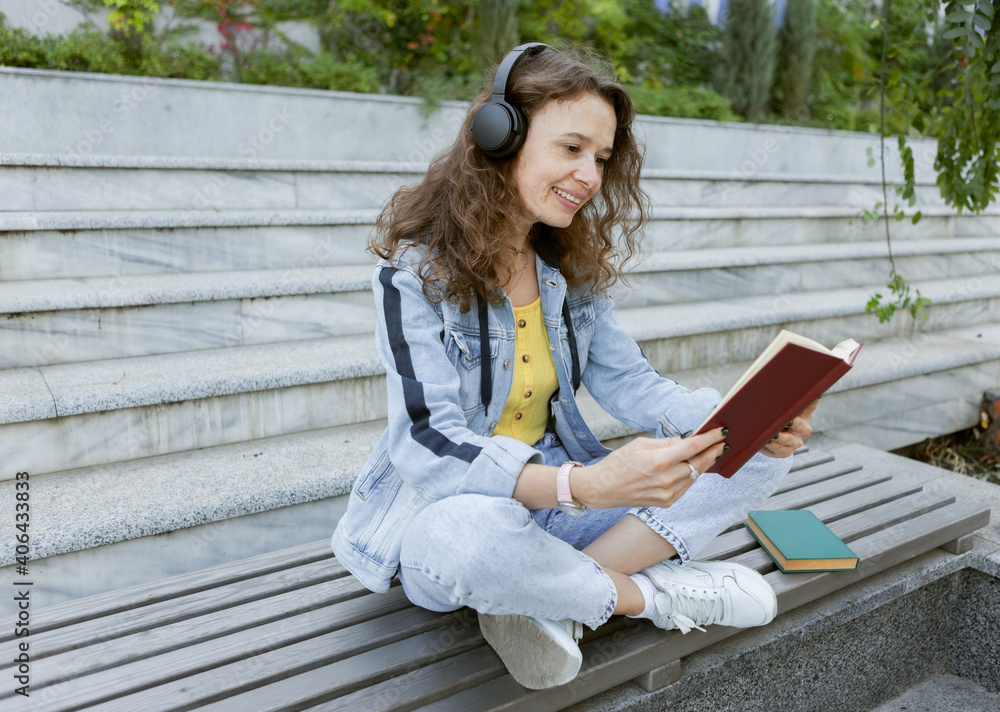 Middle-aged curly hair cheerful woman in denim clothes listening to music and reading a book while sitting on a bench on the background of stairs outdoors Middle-aged curly hair cheerful woman in denim clothes listening to music and reading a book while sitting on a bench on the background of stairs outdoors