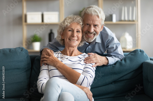 Portrait of affectionate caring middle aged man cuddling shoulders of sincere smiling mature wife relaxing on cozy couch in living room. Happy elderly married family couple enjoying weekend together.