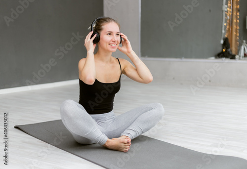 Nice cheerful woman in sportswear listening to music with headphones while sitting on mat indoor