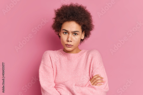 Portrait of serious angry African American woman stands with crossed hands looks irritated at camera waits for your explantions wears casual sweater isolated over pink background. Negative emotions © wayhome.studio 