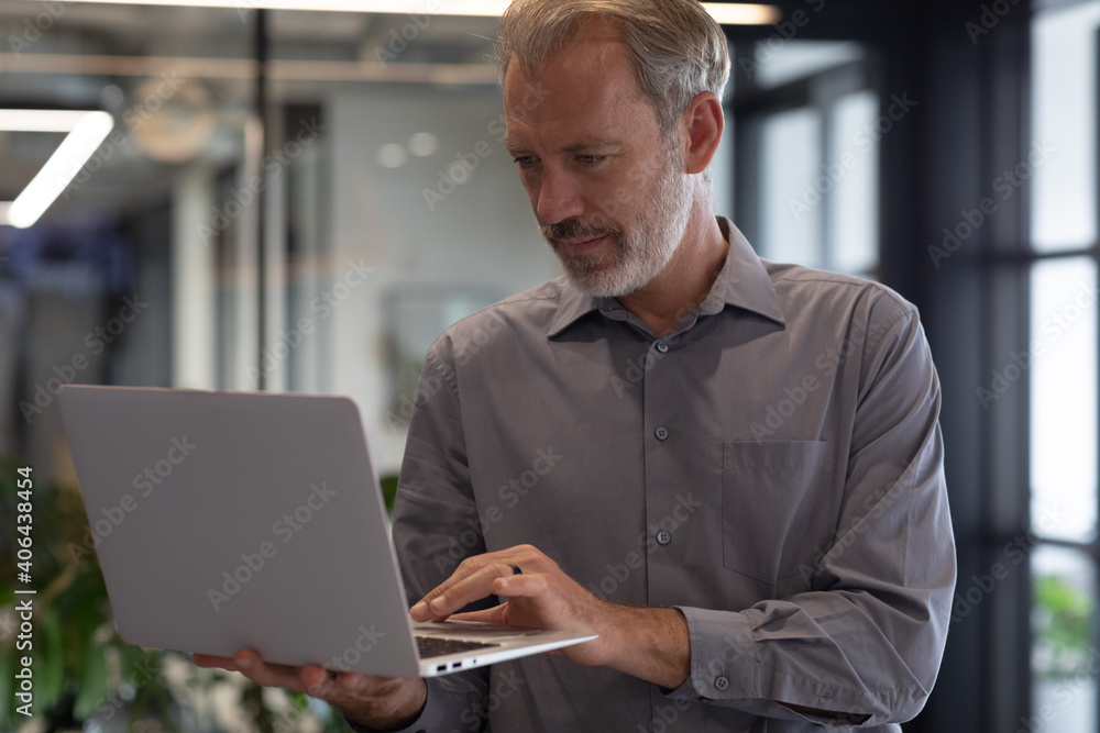 Caucasian businessman standing by window using laptop in modern office Caucasian businessman standing by window using laptop in modern office