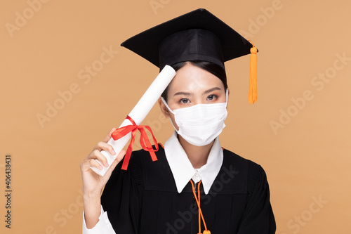 Attractive Asian woman graduated in cap and gown wear medical mask smile so proud and happiness due Virus outbrake and COVID-19 virus flu pandemic on Beige background,New normal of graduation concept photo