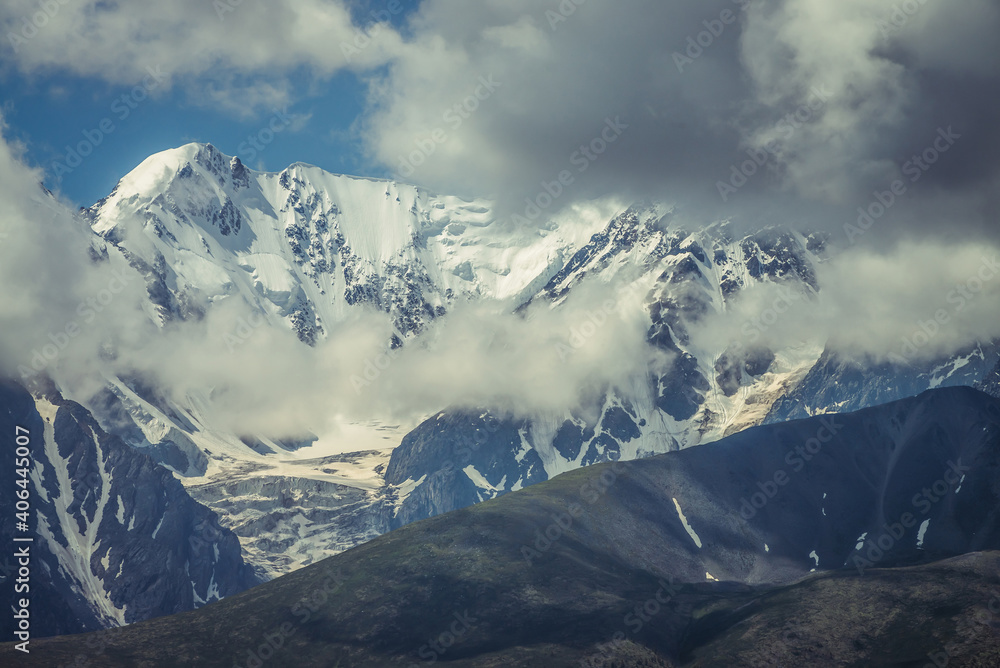 Awesome sunny mountains landscape with sunlit high snowy pinnacle among low clouds. Atmospheric highland scenery with snow-white big mountain top in sunlight. Wonderful snowy mountain range in clouds.