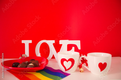 red plate in the shape of a heart with bonbons and two cups shite with red hearts and gay pride flag