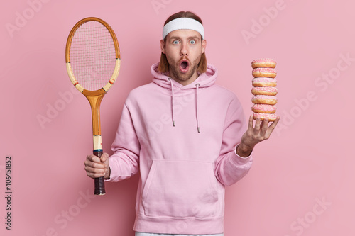 Bearded shocked hipster wears sport outfit holds tennis racket and pile of sweet doughnuts gazes surprisingly at camera with widely opened mouth has active leisure poses against pink background