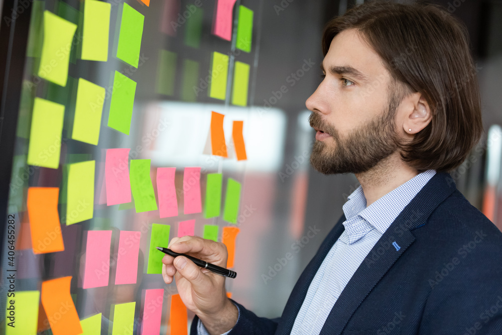 Side head shot view focused young male project manager analyzing working processes or checking progress on colored sticky paper notes on glass wall, involved in developing business strategy in office. Side head shot view focused young male project manager analyzing working processes or checking progress on colored sticky paper notes on glass wall, involved in developing business strategy in office.