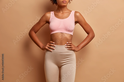 Sporty young woman has perfect body shape keeps hands on waist dressed in cropped top and leggings isolated over beige background. Health and welness concept photo