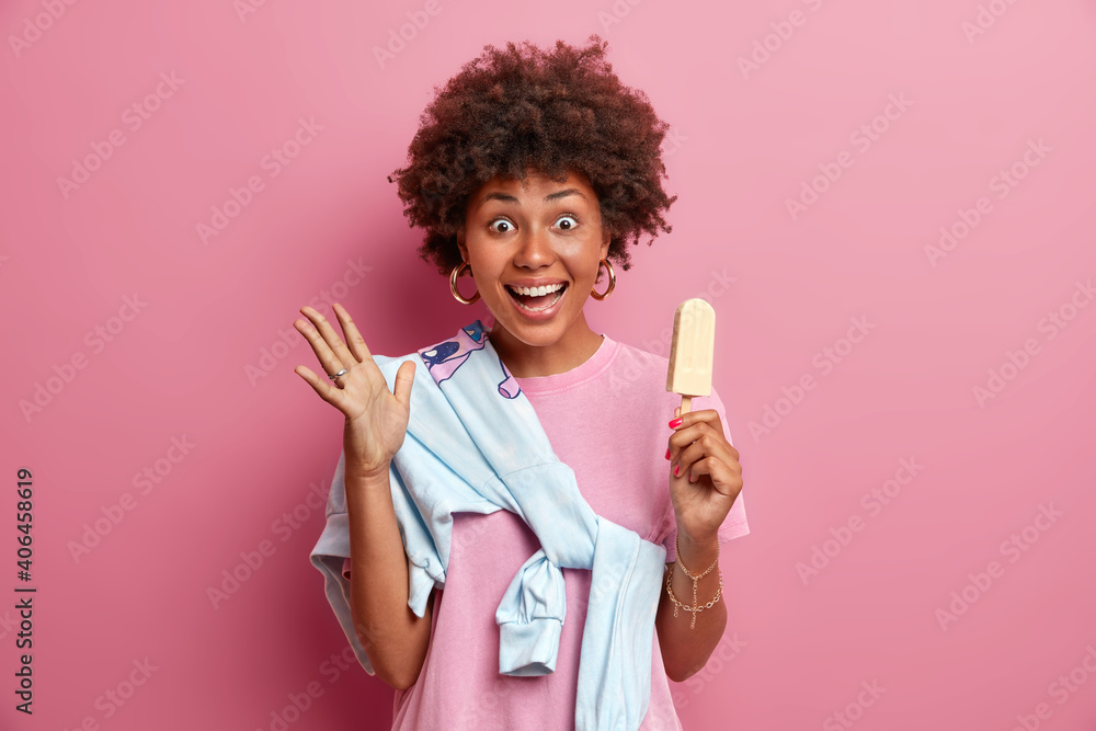 Emotional cheerful millennial girl raises hand has very glad expression holds ice cream wears sweater tied over shoulder expresses positive emotions isolated on pink wall. Woman with cold dessert Emotional cheerful millennial girl raises hand has very glad expression holds ice cream wears sweater tied over shoulder expresses positive emotions isolated on pink wall. Woman with cold dessert