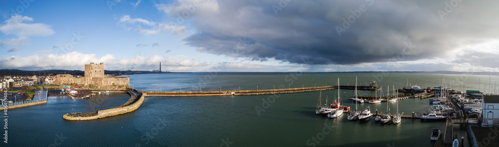 Carrickfergus near Belfast, Northern Ireland, UK. Wide aerial panorama of medieval Norman Castle, marina with yachts, harbor, breakwater and Belfast Laugh. Winter, dark stormy clouds, sunset light.