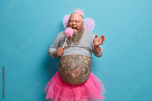 Funny bearded dad with tattooed belly entertains kids on party being holiday performer dressed like princess or fairy sings in magic wand poses against blue background. Plump man in carnival costume photo