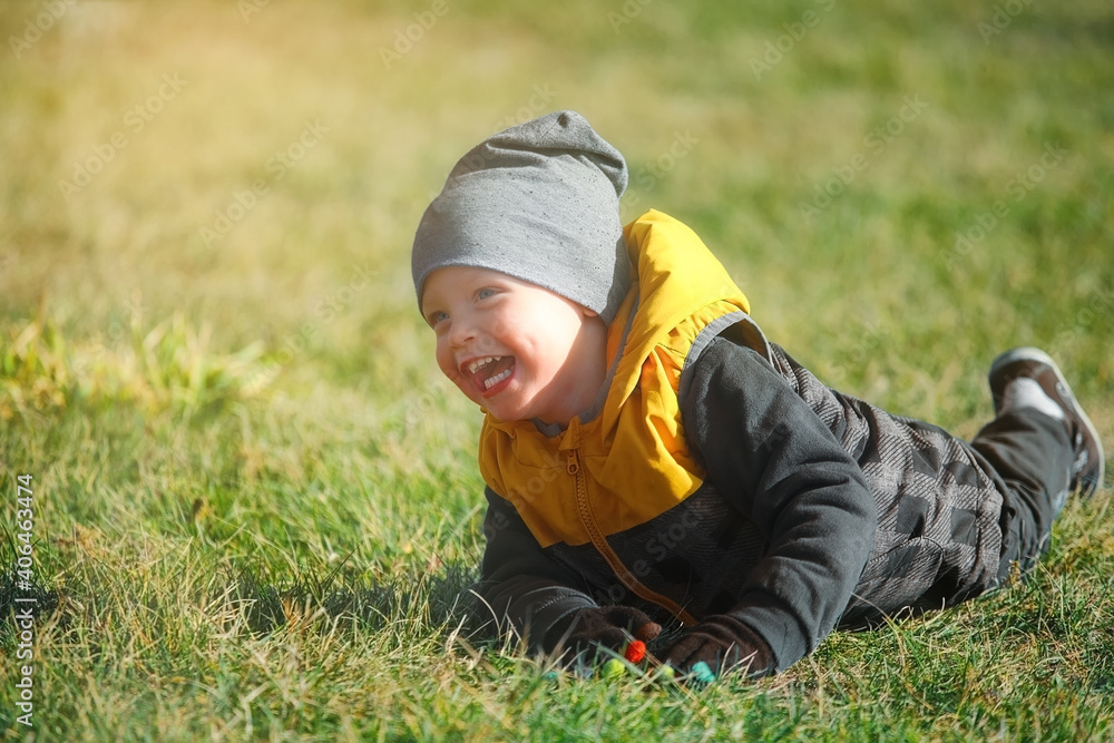 Emotional portrait of a Caucasian boy in an autumn park on a walk. A happy child in a windbreaker and hat lies on the grass and laughs. The theme of a serene childhood. Emotional portrait of a Caucasian boy in an autumn park on a walk. A happy child in a windbreaker and hat lies on the grass and laughs. The theme of a serene childhood.