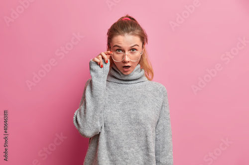 Studio shot of shocked European female model stares through transparent eyeglasses keeps mouth widely opened from astonishment cannot believe her eyes dressed in loose grey sweater isolared on pink © wayhome.studio 