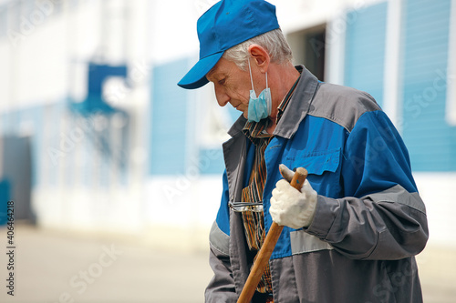 This is an elderly janitor with a broom in a medical mask on the street sweeping the territory. An old man in a work uniform works during a time of pandemic and unemployment. Close-up portrait photo