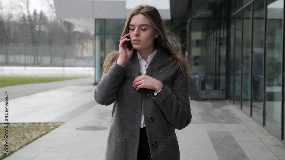 Pretty caucasian female manager businesswoman walking outside big modern business center talking on her phone feeling cold freezing. Young woman feels concerned anxious. Cold windy winder day. Pretty caucasian female manager businesswoman walking outside big modern business center talking on her phone feeling cold freezing. Young woman feels concerned anxious. Cold windy winder day.