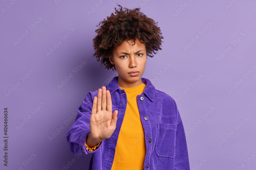 Serious annoyed dark skinned Afro American woman keeps palm in stop gesture asks not to bother her looks angrily wears purple jacket expresses restriction or denial. Do not come closer please Serious annoyed dark skinned Afro American woman keeps palm in stop gesture asks not to bother her looks angrily wears purple jacket expresses restriction or denial. Do not come closer please