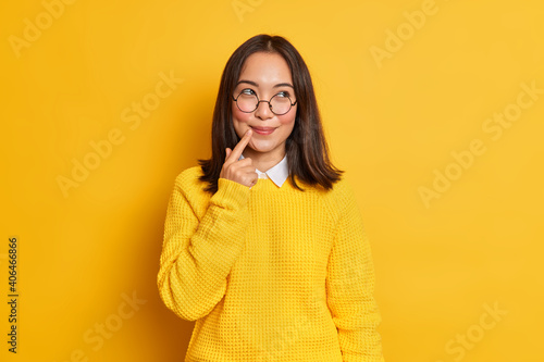 Lovely dreamy young Asian woman with dark hair keeps finger near lips wears round transparent glasses and sweater isolated over vivid yellow background recalls something funny happened with her