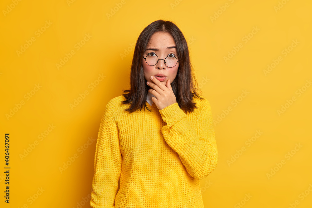 Portrait of worried surprised Asian woman holds chin looks concerned at camera wears transparent glasses and sweater isolated over yellow background. People face expressions and reaction concept Portrait of worried surprised Asian woman holds chin looks concerned at camera wears transparent glasses and sweater isolated over yellow background. People face expressions and reaction concept