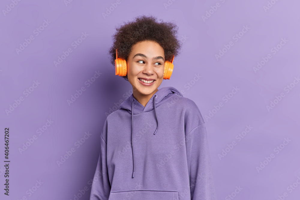 Horizontal shot of curly pretty teenage girl wears stereo headphones listens audio track looks happily aside dressed in sweatshirt isolated over purple background. Monochrome shot. Lifestyle Horizontal shot of curly pretty teenage girl wears stereo headphones listens audio track looks happily aside dressed in sweatshirt isolated over purple background. Monochrome shot. Lifestyle