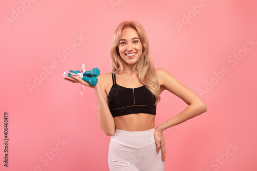 Happy fit young lady holding dumbbells tied by ribbon on pink studio background. Active lifestyle and wellness concept