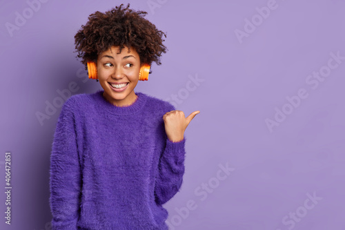 Horizontal shot of good looking cheerful African American woman listens audio track points thumb away on empty space smiles positively poses against vivid purple background advertises some item