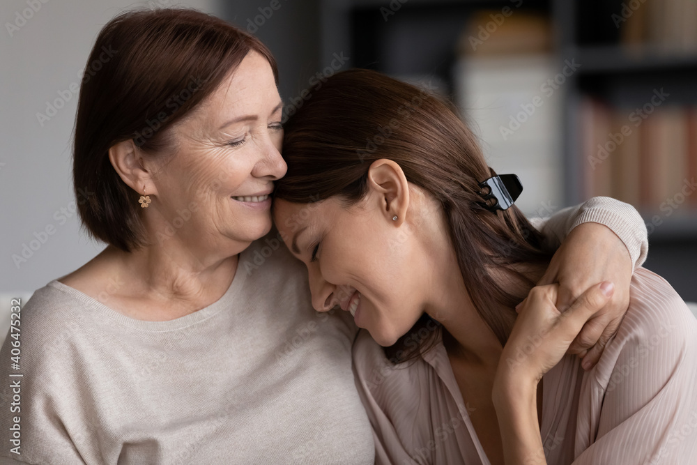 Naklejka premium Close up smiling young woman and mature mother hugging, elderly mum and grownup daughter enjoying tender moment, cuddling, holding hands, happy family spending leisure time at home together